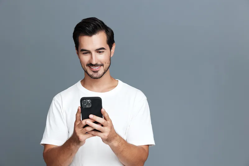 Smiling man in casual white t-shirt looking at mobile phone, standing against solid grey background. Modern lifestyle and technology concept.