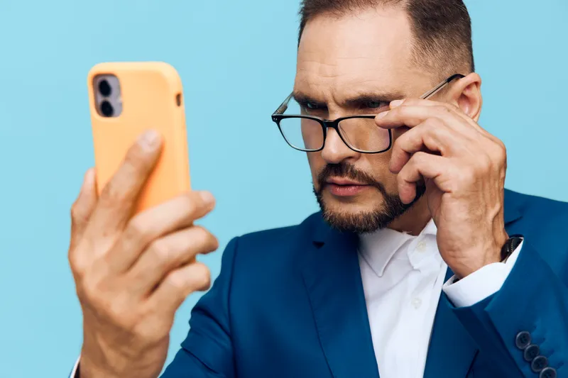 A professional man in a blue suit studies his smartphone with focused emotion. Isolated bright blue background highlights concentration and confident intent.
