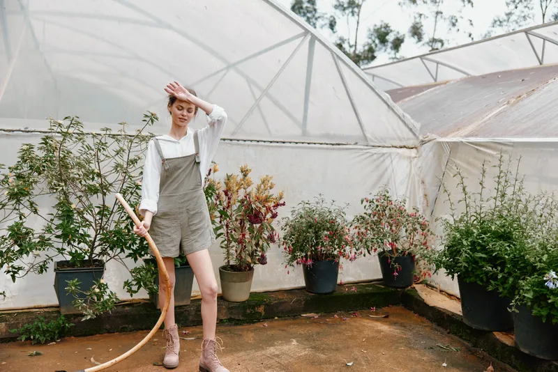 Confident woman in overalls holding a bow, standing inside a greenhouse surrounded by lush greenery and flowers, showcasing vibrant colors and seasonal beauty