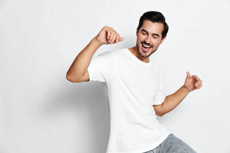 Full body smiling cheerful young man wearing white t-shirt dancing happily with joy against a plain solid background conveying a sense of freedom and happiness.