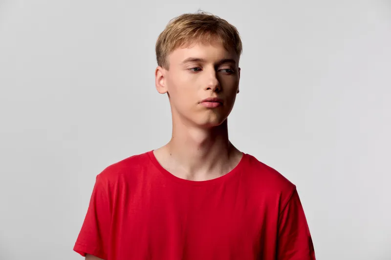 Young man in a red shirt stands against a neutral studio backdrop with calm expression, clean lighting, and a minimalist mood, suitable for fashion, lifestyle and product promotion