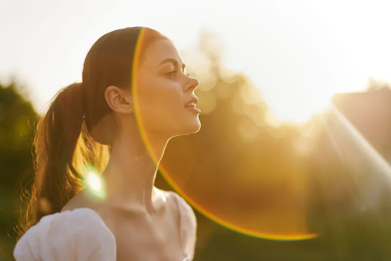 Young woman enjoying sunlight with a peaceful expression, dressed in a white blouse, surrounded by nature, evoking a sense of tranquility and warmth