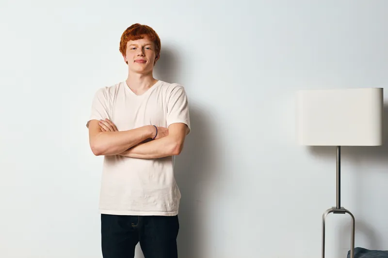 Young man with red hair standing with arms crossed near wall and lamp. Casual style, confident expression, indoor portrait with simple background and relaxed pose.