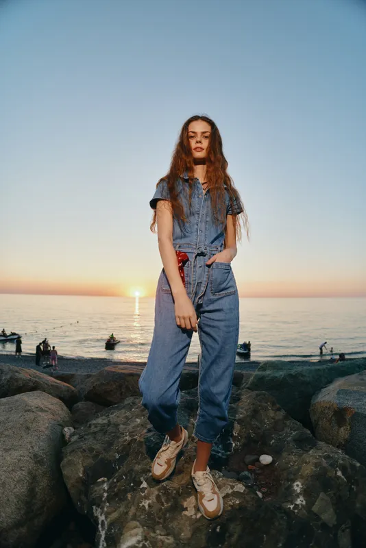 young woman in denim outfit standing on rocks by the sea at sunset with long hair and casual shoes under clear sky