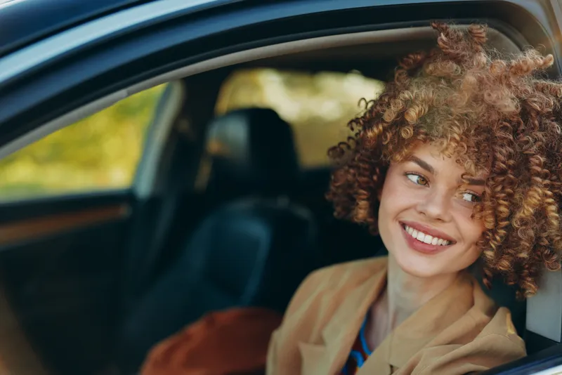 Smiling woman with curly hair sitting in a car, wearing a casual blazer, natural light highlights her cheerful smile. Authentic lifestyle moment that conveys relaxed travel