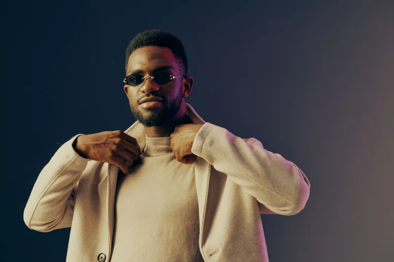 Stylish young Black man adjusting his coat with sunglasses, set against a gradient background, conveying confidence and sophistication in modern fashion