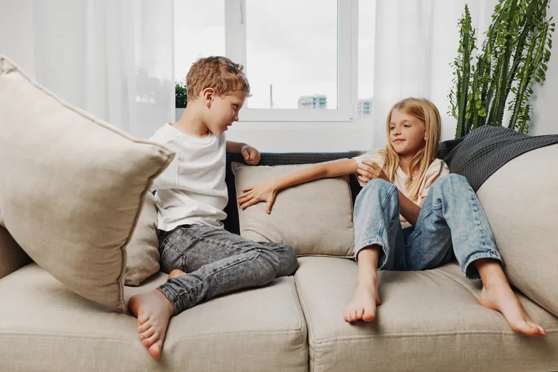 Two barefoot children sitting on beige sofa in casual clothes, playing and interacting in bright living room with large window and plants.
