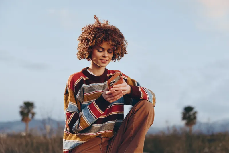 Woman lifestyle outdoors, smiling with curly hair, holding smartphone, phone in hands, wearing cozy sweater and brown pants, relaxed pose, soft light, autumn landscape background, candid
