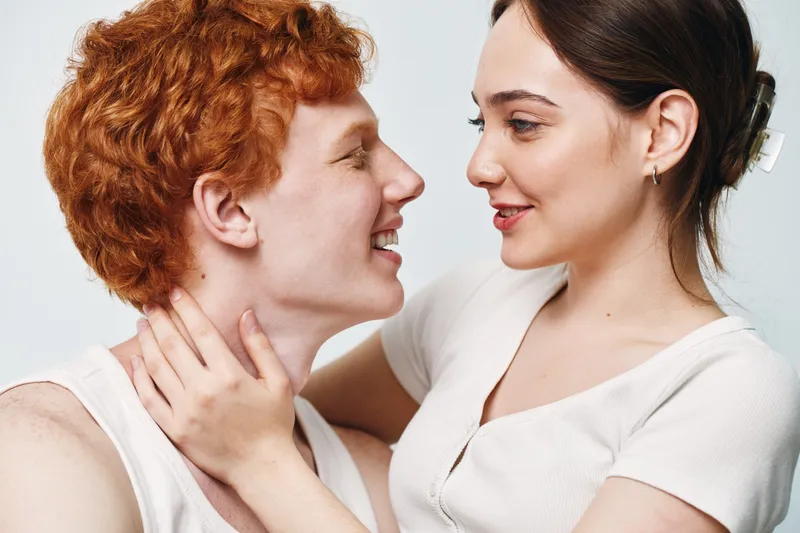 Young couple facing each other with smiles, closeup of affectionate moment. Woman holding man by neck, positive emotion and intimacy in casual clothing on white background.