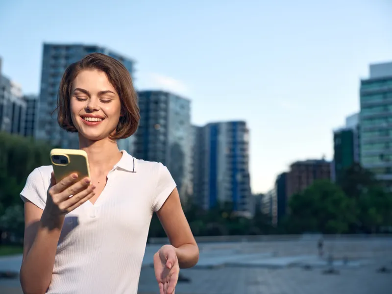 A cheerful professional woman stands outdoors in a modern city setting, smiling while looking at her smartphone. She projects confidence and positive energy under daylight.