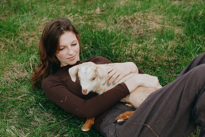 A goat and kid cuddle with a woman outdoors, a lifestyle moment of bonding and relaxation in a meadow. A tender, calm scene of companionship, nature, and simple joy in bright daylight.