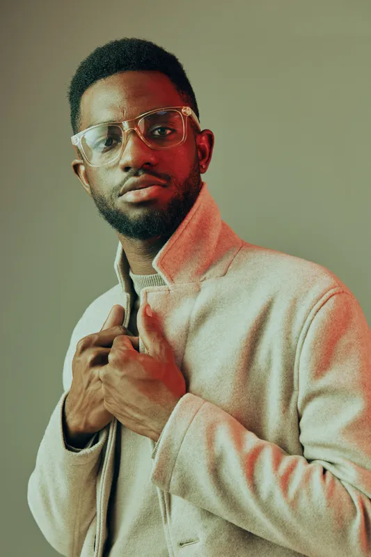 Stylish young African American man in a trendy beige jacket, displaying confidence with modern glasses against a warm backdrop, ideal for lifestyle contexts