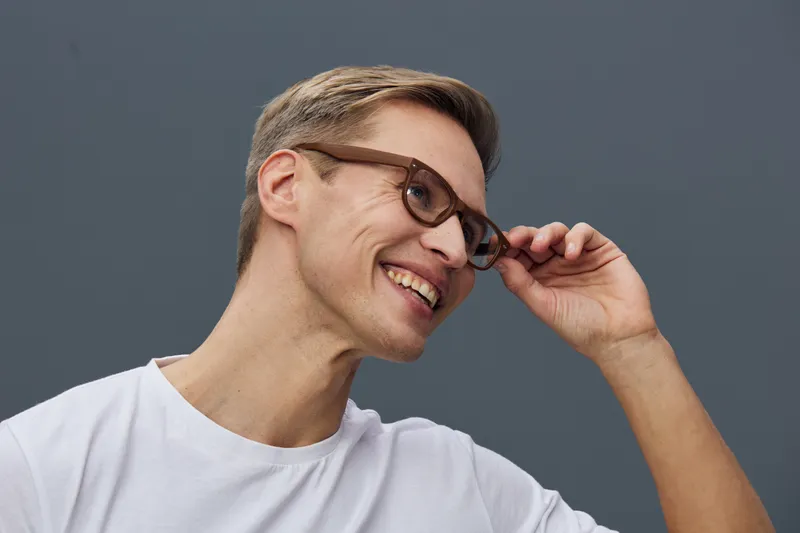 Smiling man with glasses, young adult, casual white t-shirt, cheerful expression, modern appearance, isolated on gray background, studio portrait, happy mood.