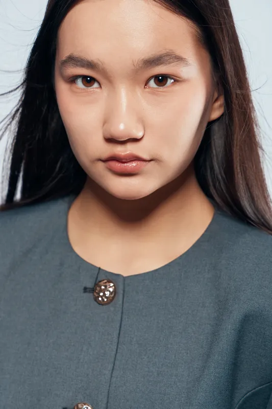 Portrait of young Asian woman with long dark hair wearing grey jacket with decorative buttons, looking seriously at camera with neutral expression on white background in studio.