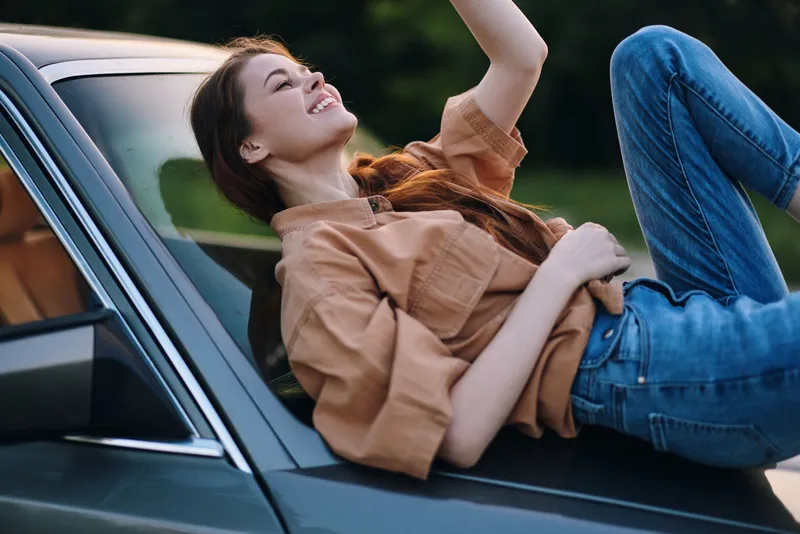 Happy young woman relaxing on car hood in casual clothes, enjoying summer day in nature, with joyful expression and vibrant greenery in background.