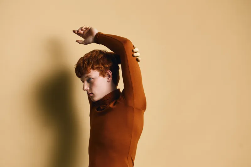 Young man with curly red hair wearing brown turtleneck stretching arms against beige background, side view, casual fashion and relaxed pose in studio lighting.