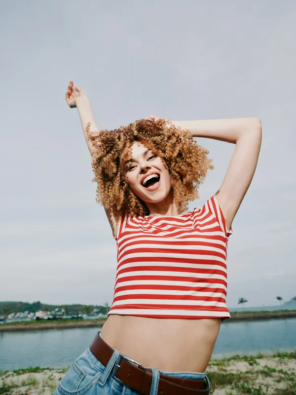 Joyful young woman dances outdoors wearing a red and white striped crop top, denim jeans, and a carefree smile against a bright sky and calm lake backdrop in a peaceful morning light.