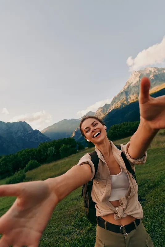 Young brunette hiker with backpack takes a cheerful selfie in a sunlit mountain meadow, extending her arms toward the camera with a bright smile.