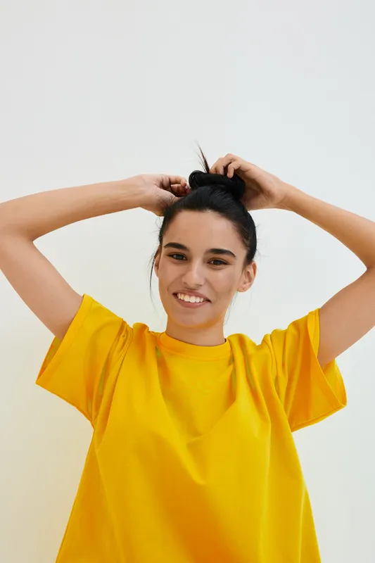 smiling woman with black hair wearing bright yellow t-shirt, fixing hair bun against plain white background, cheerful expression and casual style