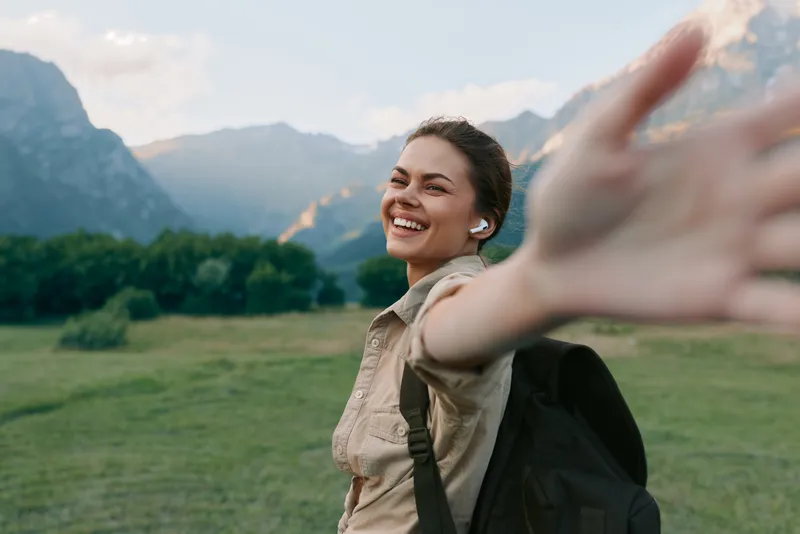 Young woman with dark hair wearing earbuds and a backpack smiles joyfully while reaching out her hand in a green mountain valley during a sunny day.