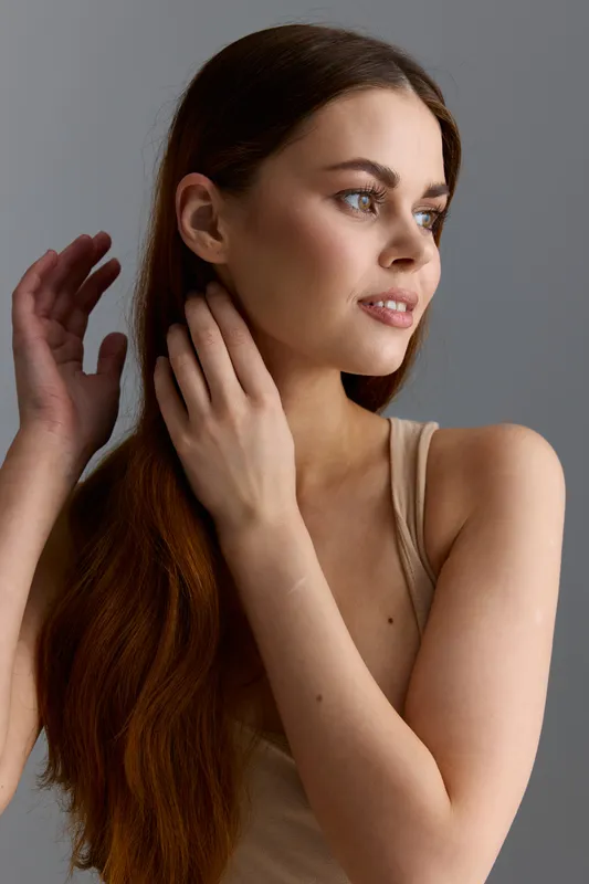 Young woman with long brown hair wearing a beige tank top, looking thoughtfully to the side with soft lighting and neutral studio background.
