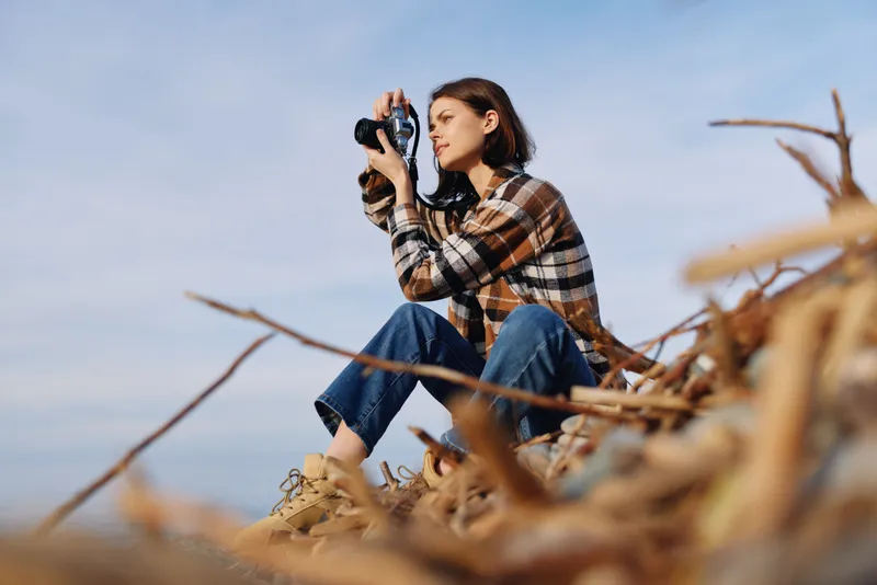 Female photographer in casual plaid shirt sits on dry reeds with a camera, gazing through the lens against an open sky, capturing nature and candid moments outdoors.
