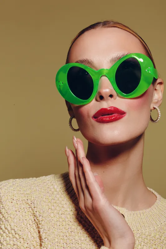 Stylish woman with green oversized sunglasses and red lipstick poses against beige background. Fashion portrait with trendy accessories and confident expression in studio lighting.