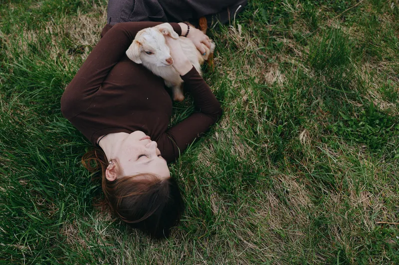 Relaxed woman outdoors lying on grass, cuddling a goat kid, sunlit moment, peaceful bond, casual clothing, natural colors, fresh air, rural setting, candid lifestyle