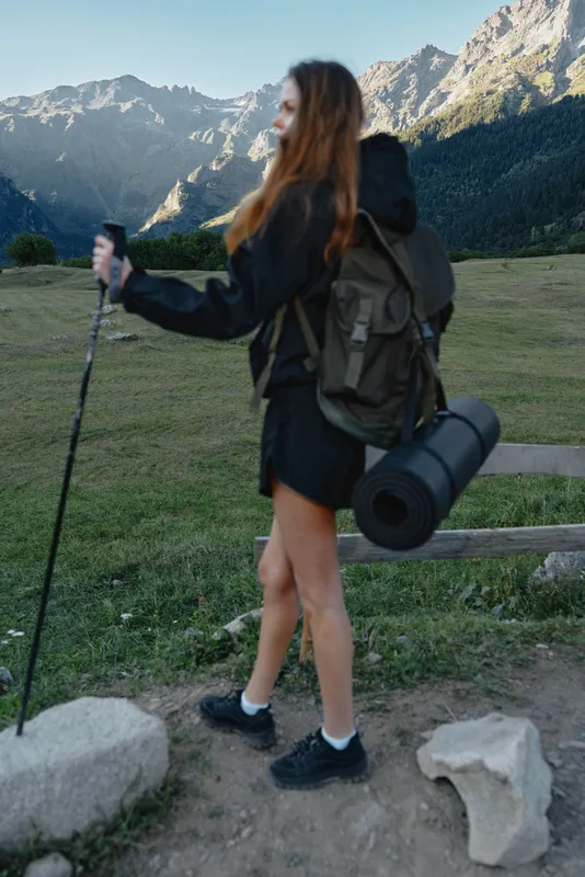 A female hiker with a backpack stands on a rocky trail, gazing toward alpine peaks. The scene conveys outdoor adventure, freedom, and exploration in a wide, natural landscape.