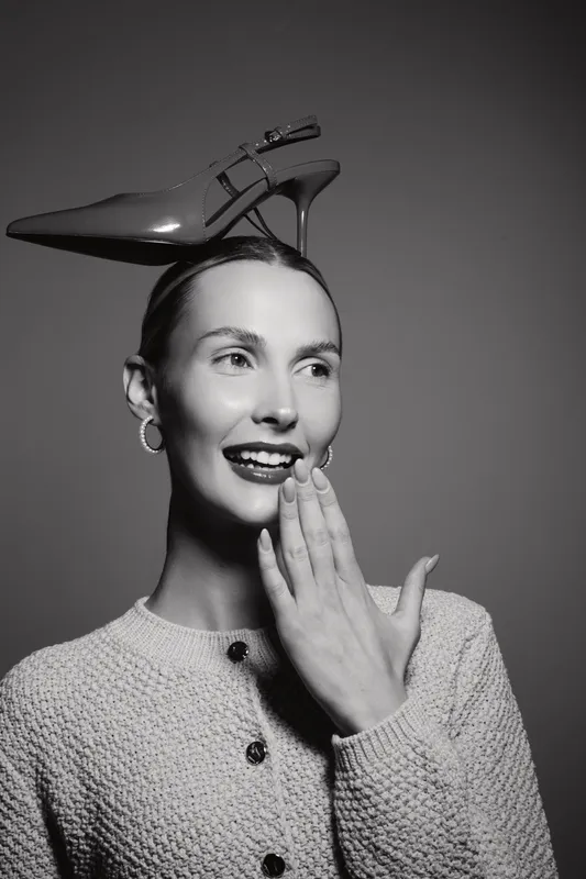Young caucasian woman with fair skin smiling and holding hand near mouth with high heel shoe on head black and white.