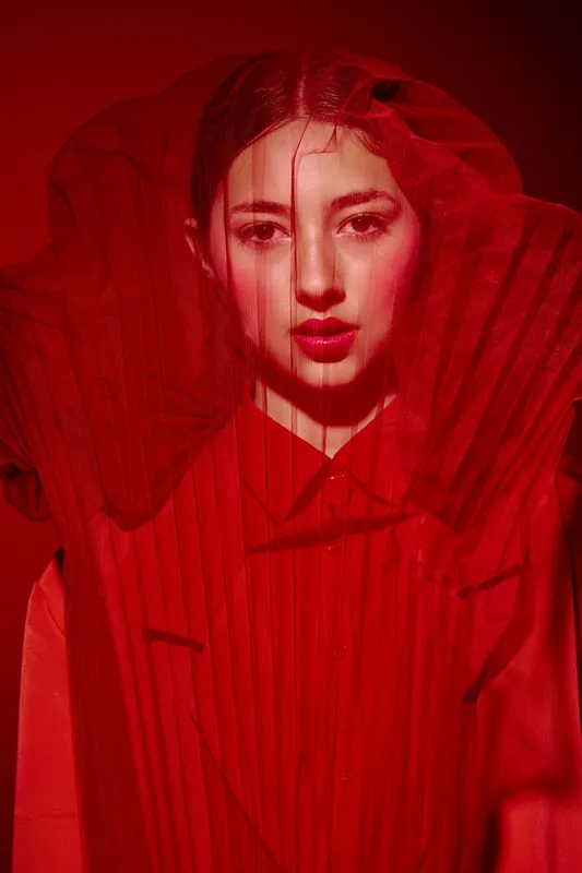 Portrait of a young woman in vibrant red fashion under sheer fabric with intense gaze and bold makeup. Studio shot with creative lighting emphasizes beauty and fashion theme.