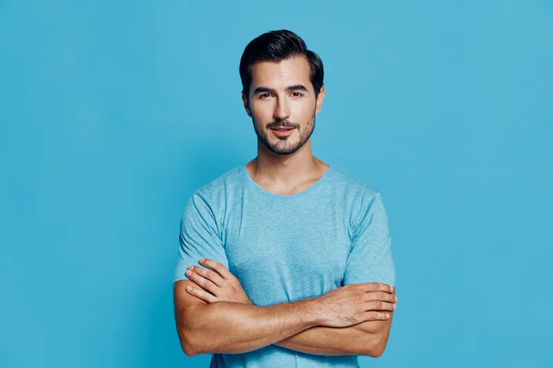 Handsome confident young man wearing a light blue t-shirt stands with arms crossed, smiling at the camera against a bright turquoise background. People lifestyle concept.