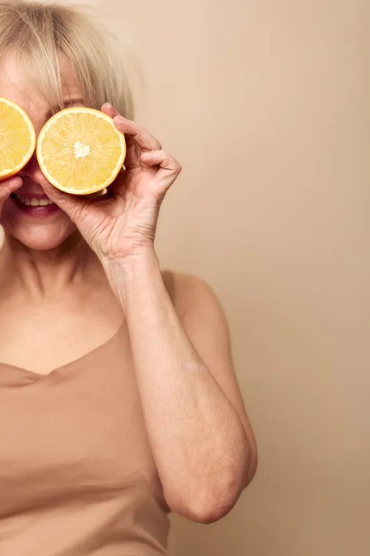 Smiling mature woman holds two halves of fresh orange in front of eyes on beige background. Concept of healthy lifestyle, vitamin c, natural beauty and joyful senior female wellness.