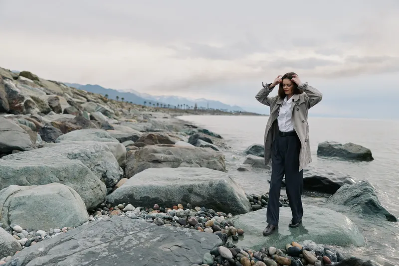 Young woman in trench coat stands on rocky beach near calm sea under cloudy sky. Concept of autumn fashion, nature, and contemplative mood by the water shore.