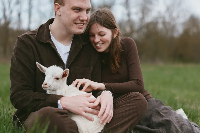 Happy couple outdoors with a white goat kid, gentle interaction, warm smiles, natural light, intimate moment on a grassy field, casual clothing, lifestyle portrait highlighting connection between