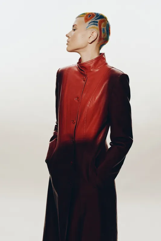 Stylish woman with colorful short hair wearing a red leather coat stands in profile pose against plain bright background, showcasing modern fashion and bold hairstyle.