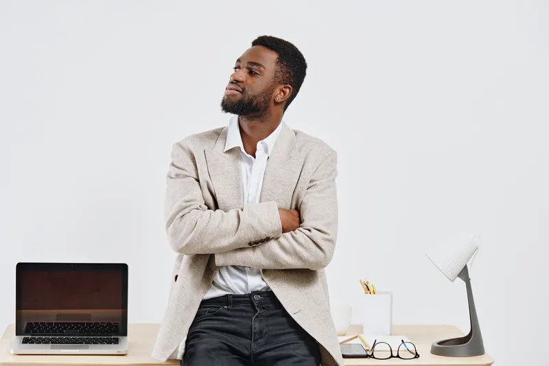 Confident young man with beard dressed in casual blazer leaning on modern office desk with laptop and accessories isolated on white background. Business casual lifestyle concept