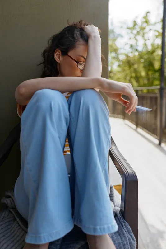 Casual woman at home, sitting on a balcony with relaxed posture, blue jeans, glasses, soft light, candid lifestyle moment, contemplative mood, gentle breeze, urban greenery backdrop