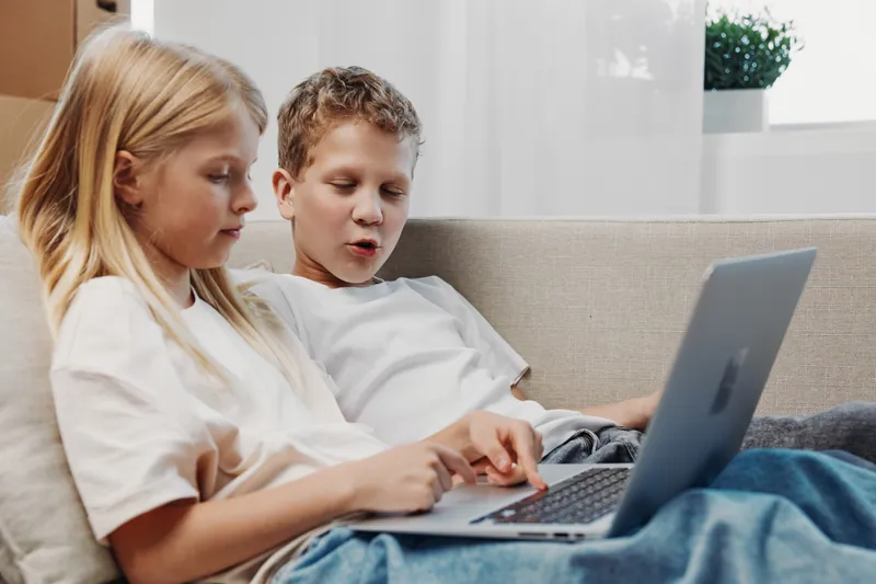 Two children sitting on a couch using a laptop together in a bright room. Concept for technology, learning, and childhood interaction.