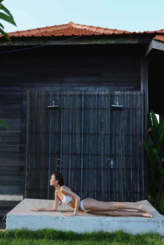 A young woman in white swimwear practices yoga on a stone platform outdoors near a wooden house with a bamboo door. The scene captures a calm lifestyle and wellness concept.