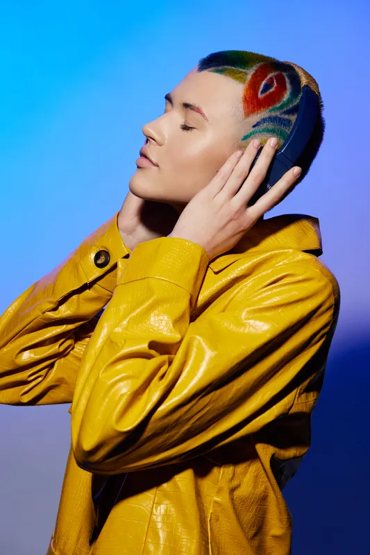 Young woman with colorful hair art and vibrant yellow jacket posing thoughtfully against blue background, showcasing modern style and creative self-expression.