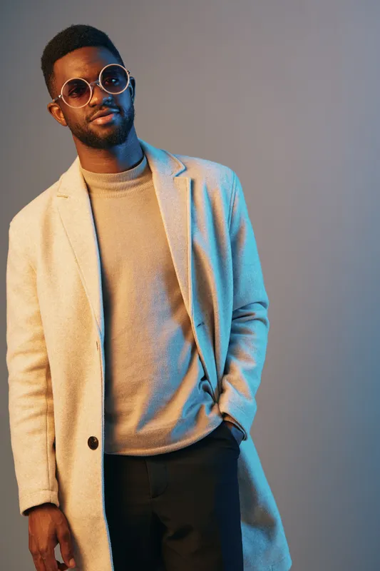 Stylish man in sunglasses wearing a beige coat and sweater against a modern blue background, showcasing contemporary fashion and confidence