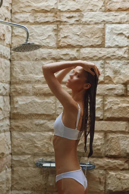 woman showering outdoors in white underwear with stone wall background, wet hair, relaxed, natural light, bathing, summer wellness, fresh water spray