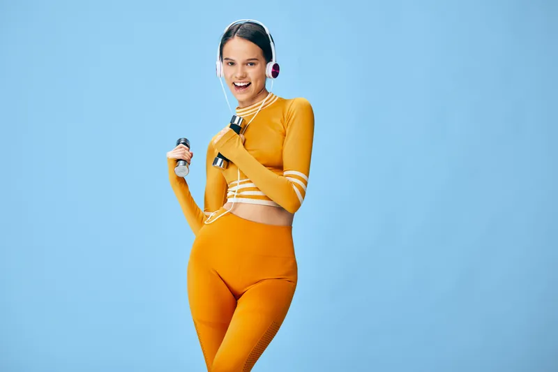 Woman in bright orange athletic wear strikes a dynamic pose with headphones and a bottle against a blue backdrop, exuding energetic fitness style, movement and a healthy lifestyle for sport