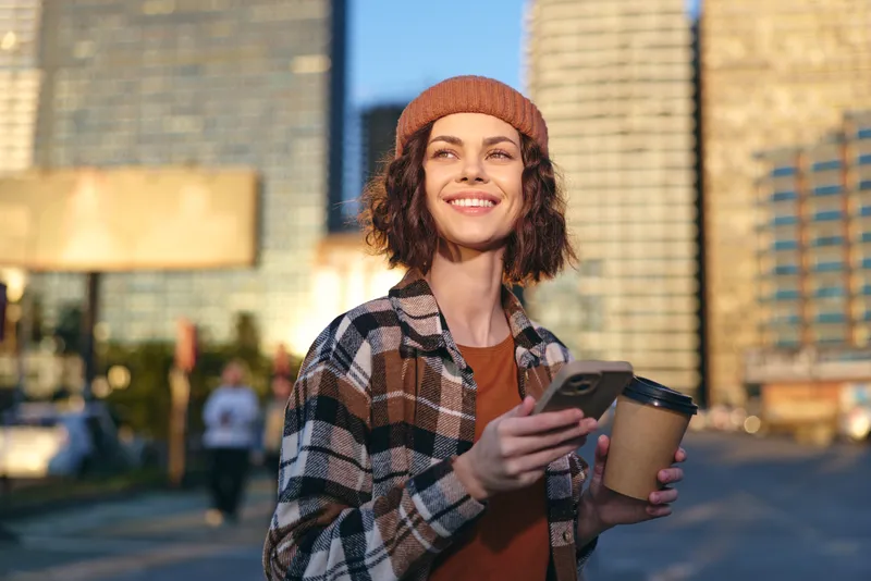 woman coffee phone smile urban beanie sunlight candid Young woman in a beanie holds coffee and phone on a city street, smiling in golden hour glow; authenticity, mindful living and emotional