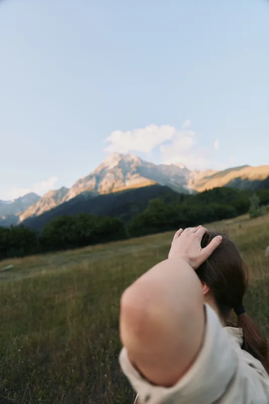 A young woman in a white shirt stands in a mountain meadow with her hand behind her head, gazing toward distant peaks.