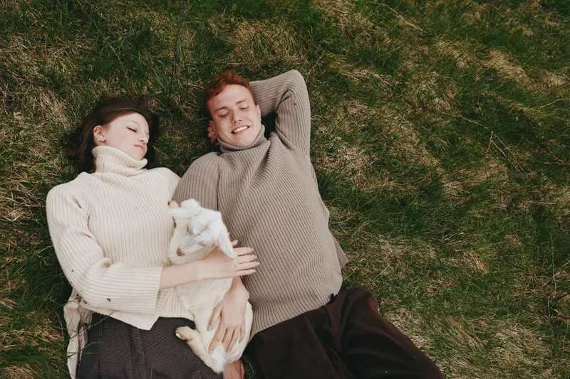 A young couple lies on the grass with a baby goat in their arms, enjoying a relaxed lifestyle moment in cozy sweaters during a calm outdoor day.