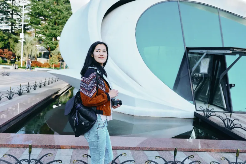 Traveler with backpack beside futuristic building, urban exploration scene, casual fashion, architecture details, reflective glass, morning light, photography gear in hand