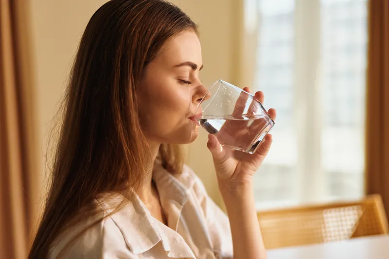 Woman drinking water from a clear glass at home in the morning, portrait of a relaxed young adult hydrating with natural light, casual clothing and calm domestic interior scene.