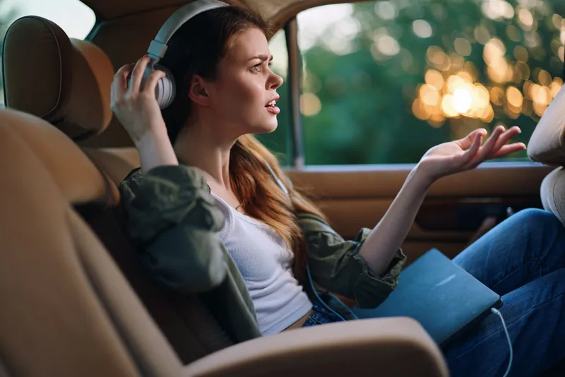 Young woman listening to music in a car during sunset, expressing emotions, wearing casual clothes, with a serene nature background.